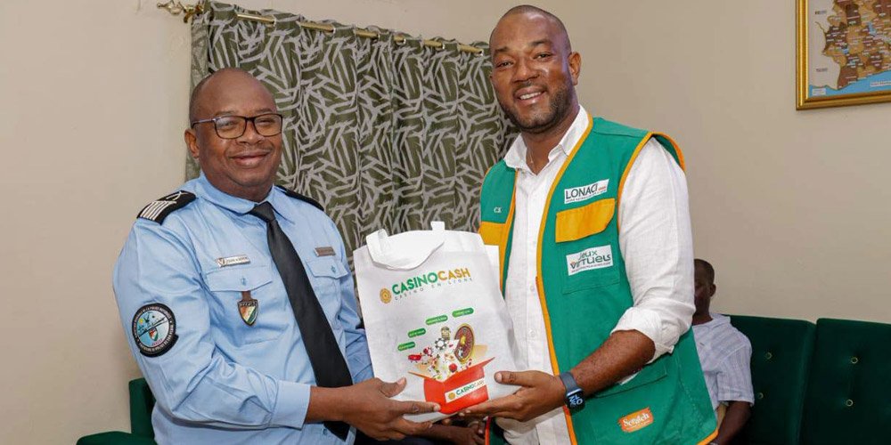 Two men exchange a white bag labeled 'CASINO CASH' while smiling; one wears a light blue security/uniform shirt, the other a green-yellow vest in a room with a patterned curtain.