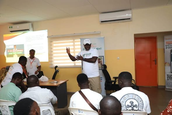 find more africa #findmoreafrica › Trainer in white shirt and cap speaks to a seated group in a classroom; projector screen and banners visible in a training session hall.