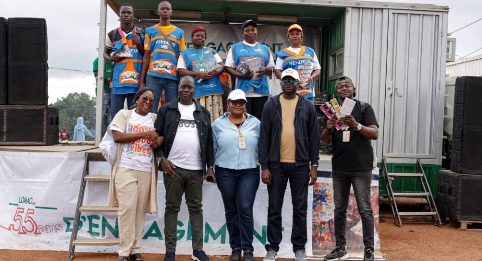 Nine people pose on a makeshift stage built from a shipping container, some wearing blue vests and holding flyers during a community event.
