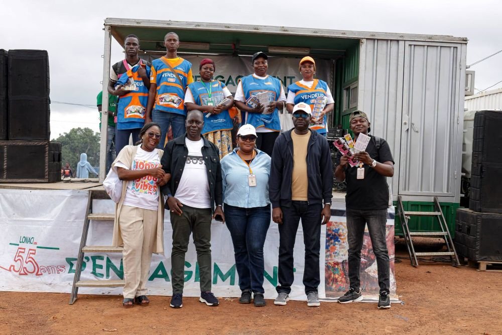Nine people pose on a makeshift stage built from a shipping container, some wearing blue vests and holding flyers during a community event.