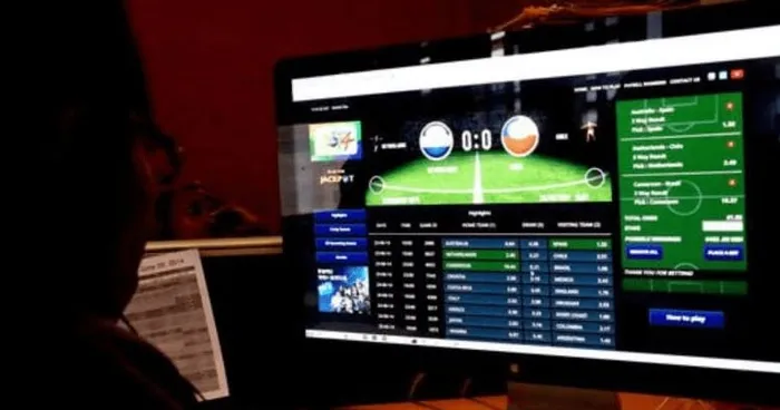Person viewing a computer monitor displaying a soccer match betting dashboard with a green field and score 0:0