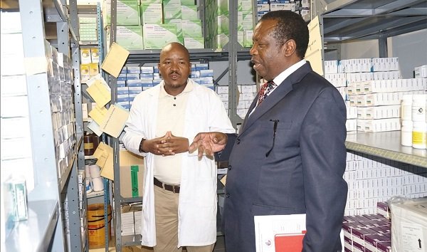 Two men in a storage room discuss inventory; one in a lab coat, the other in a suit holding a folder near shelves of boxes and supplies in a warehouse.