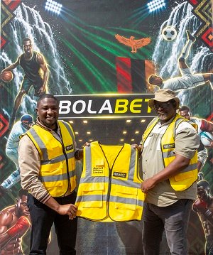 Two men in yellow safety vests pose on a BolaBet stage, presenting a high-visibility vest in front of a sports-themed backdrop.