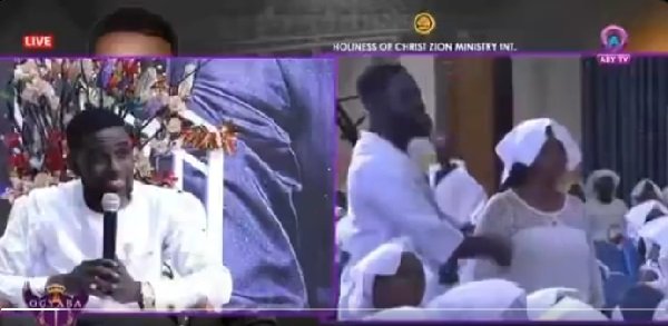 find more africa #findmoreafrica › Live church service scene: a man in a white suit speaks into a microphone while attendees in white garments sit and kneel in the background on a broadcast set.