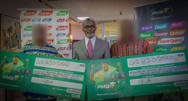 Three men hold oversized green checks at a prize ceremony; center man in a suit with a pink tie, others blurred, banners in the background.] - image text trimmed?