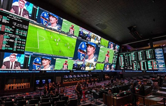 Large wall of video screens showing live sports events in a betting lounge with spectators nearby.