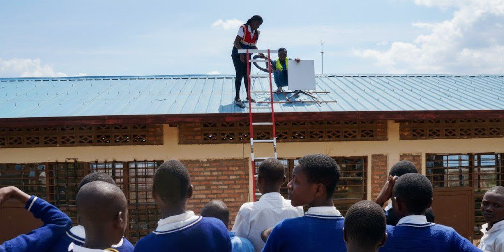 Two workers in safety vests on a blue metal roof repair or install equipment, with a tall ladder and students watching from below a school building.
