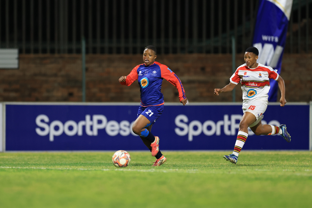 Soccer players chase the ball on a grass field during a night match, one in blue-red kit and another in white-red kit behind him.