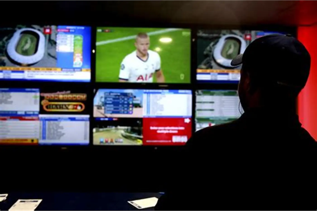 Silhouette of a person standing in front of a wall of monitors displaying sports feeds and data panels.