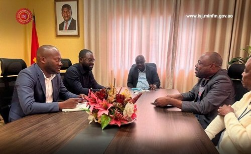 Five men sit around a conference table in a formal meeting room; a flower arrangement sits in the center, with a flag, a framed portrait, and draped windows in the background. The website www.isi-minfin.gov.ao is visible in the upper-right.