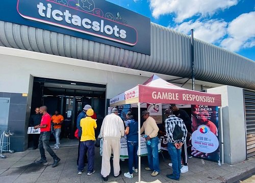 People queue at a 'GAMBLE RESPONSIBLY' booth outside a building with a 'tictacslots' sign.