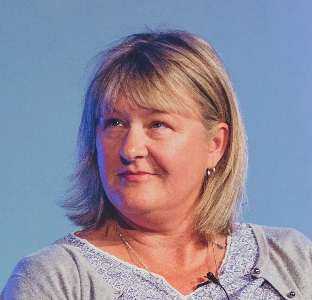 Close-up portrait of a smiling middle-aged woman with shoulder-length blond hair against a blue background, wearing a light patterned top and a necklace.