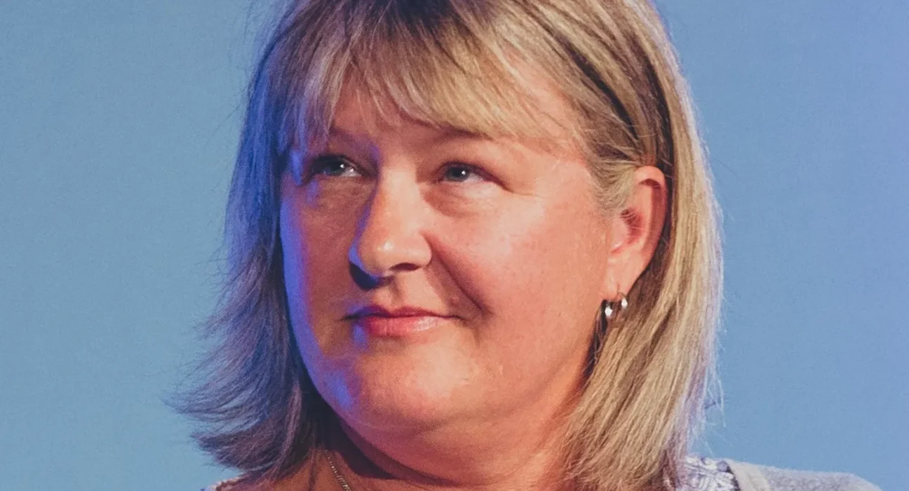 Close-up portrait of a smiling middle-aged woman with shoulder-length blond hair against a blue background, wearing a light patterned top and a necklace.