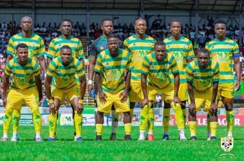A soccer team in yellow and green uniforms posing for a pregame photo on a grass field with stands in the background.