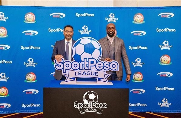 Two men in suits pose beside a large soccer-ball sign that says SportPesa League, with a blue branded backdrop behind them.