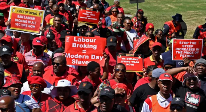 Large crowd of protesters wearing red, holding placards with messages about labour, living costs, and wages, including'Labour Breaking = Modern Day Slavery.'