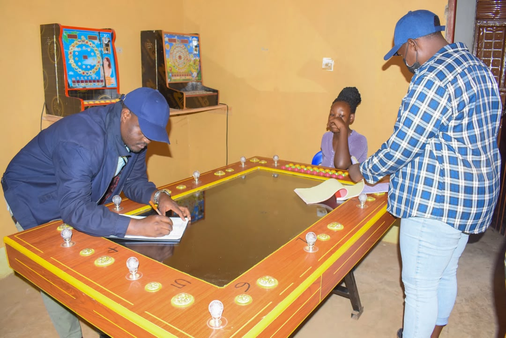 Two men and a girl sit around a casino-style gaming table; one man signs a clipboard while the others watch, arcade machines in the background.