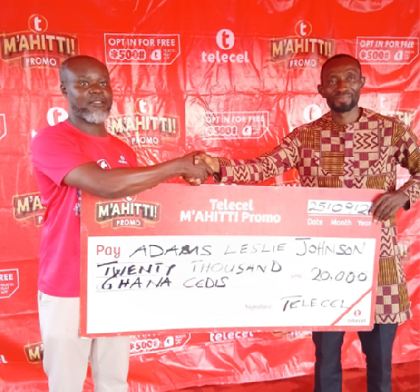 Two men shake hands while holding a large ceremonial cheque in front of a red promotional backdrop. Banners read promotional branding in the background.