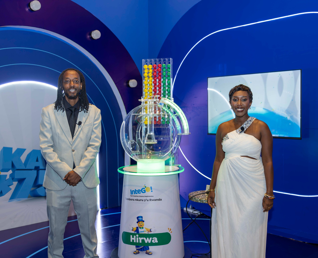 Two presenters stand at a blue exhibition booth next to a transparent lottery-style machine filled with yellow, red, green, and blue balls.